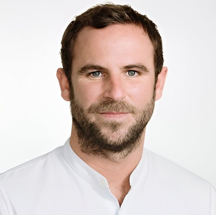 Man with a beard and short hair wearing a white shirt against a plain background.